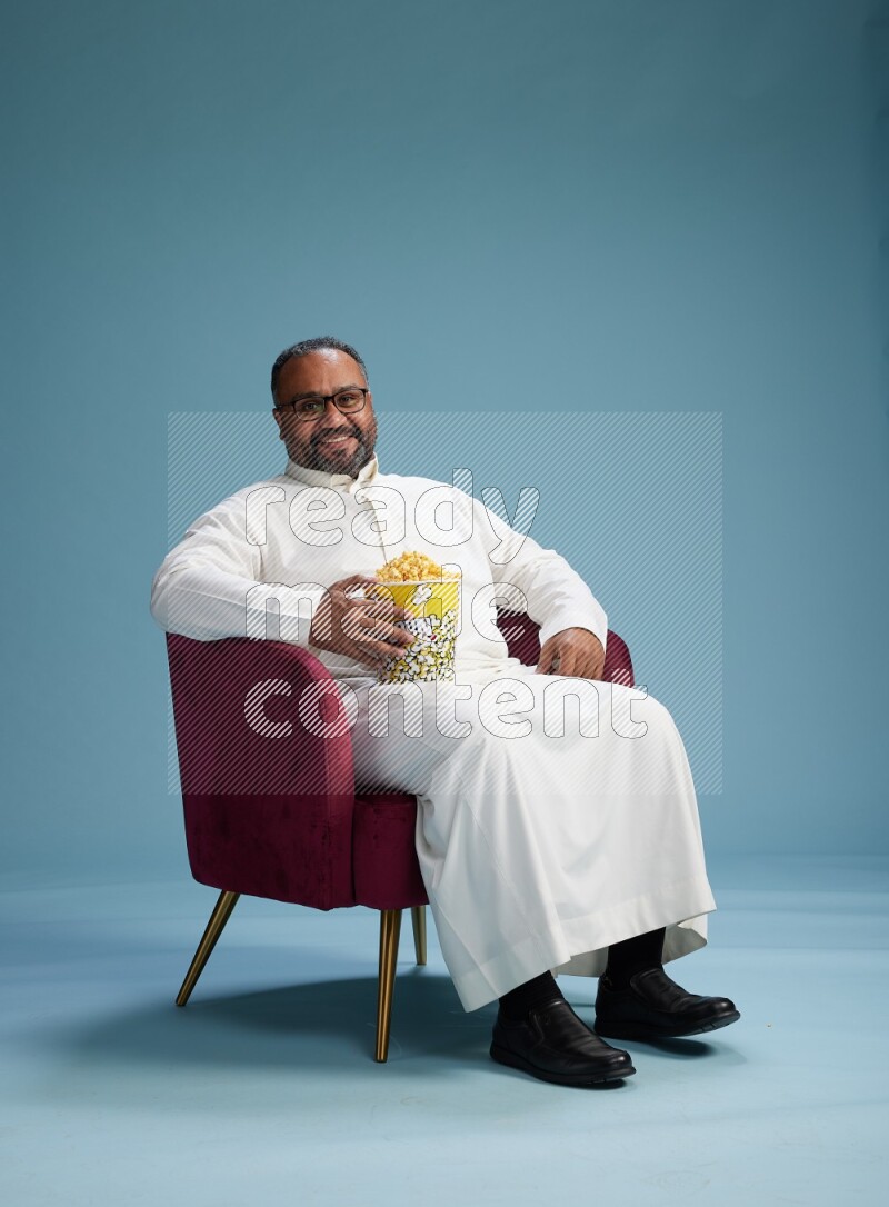 Saudi Man without shimag sitting on chair eating popcorn on blue background