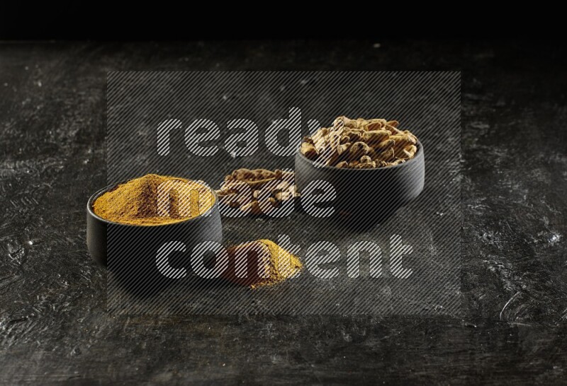 2 pottery black bowls, one full of turmeric powder and the other full of dried turmeric whole fingers on a textured black flooring
