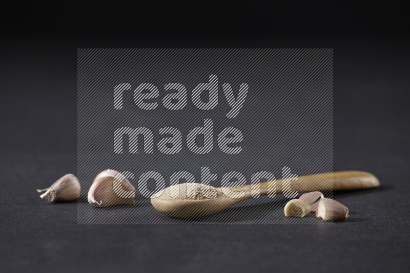 A wooden spoon full of garlic powder with cloves beside it on a black flooring