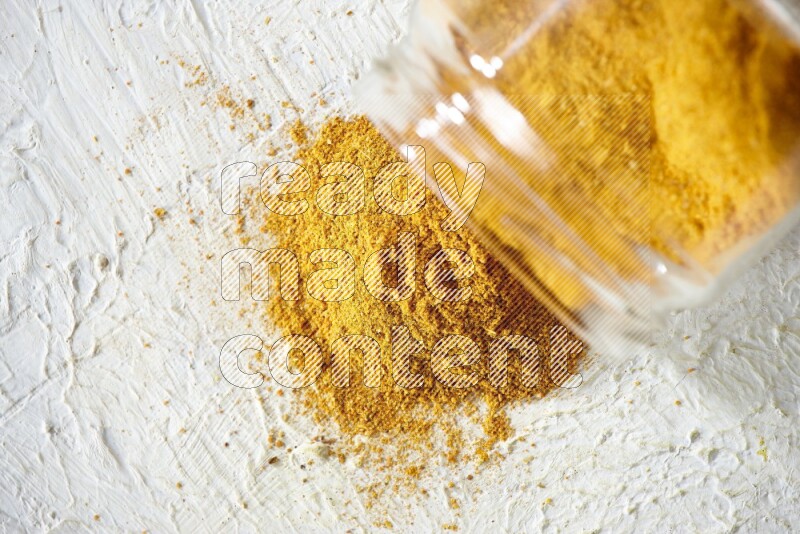 A flipped glass spice jar full of turmeric powder and powder fell out it on textured white flooring