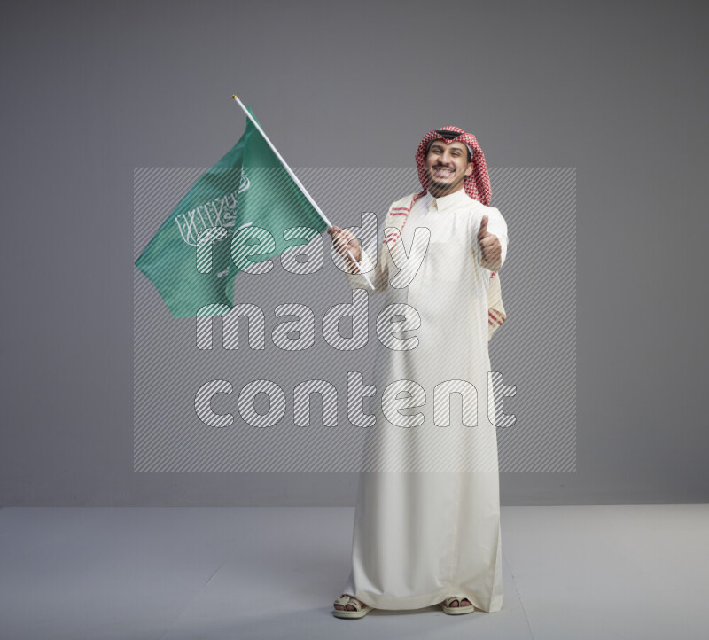 A Saudi man standing wearing thob and red shomag raising big Saudi flag on gray background