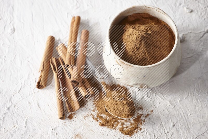 Ceramic beige bowl full of cinnamon powder and a metal spoon with cinnamon sticks next of it on a textured white background