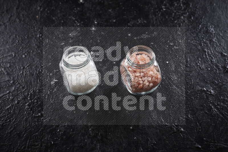 2 glass jars one is filled with coarse himalayan salt and the other with coarse sea salt on black background