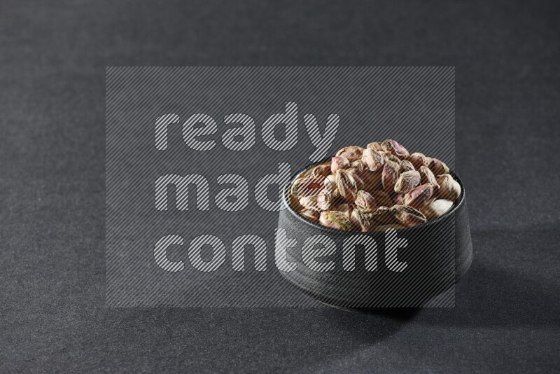 A black pottery bowl full of peeled pistachios on a black background in different angles
