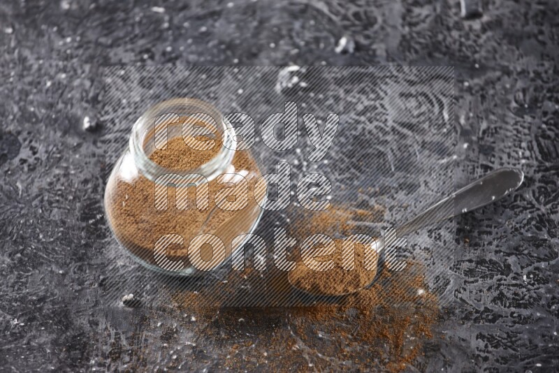Herbal glass jar full of cinnamon powder and a metal spoon full of powder on textured black background