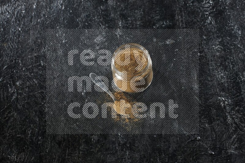 A glass jar and a metal spoon full of cumin powder on a textured black flooring