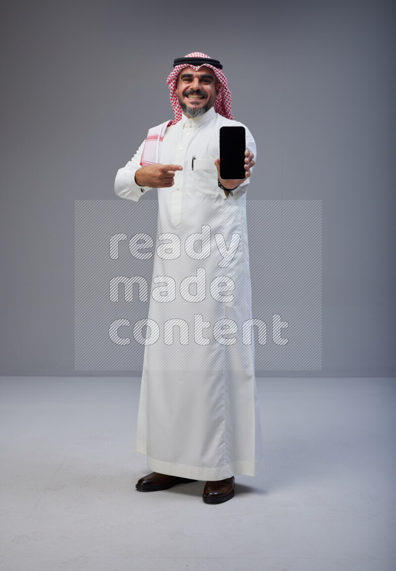 Saudi man Wearing Thob and red Shomag standing showing phone to camera on Gray background