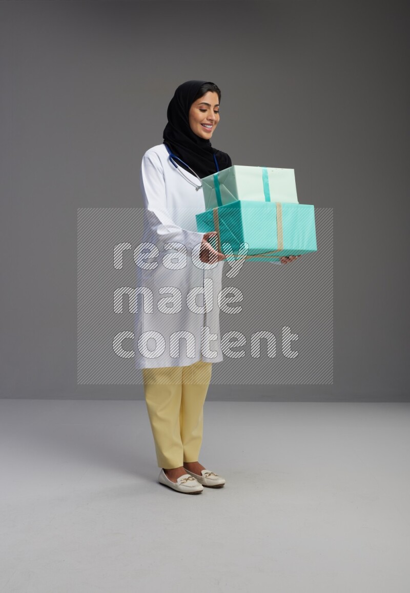 Saudi woman wearing lab coat with stethoscope standing holding gift box on Gray background