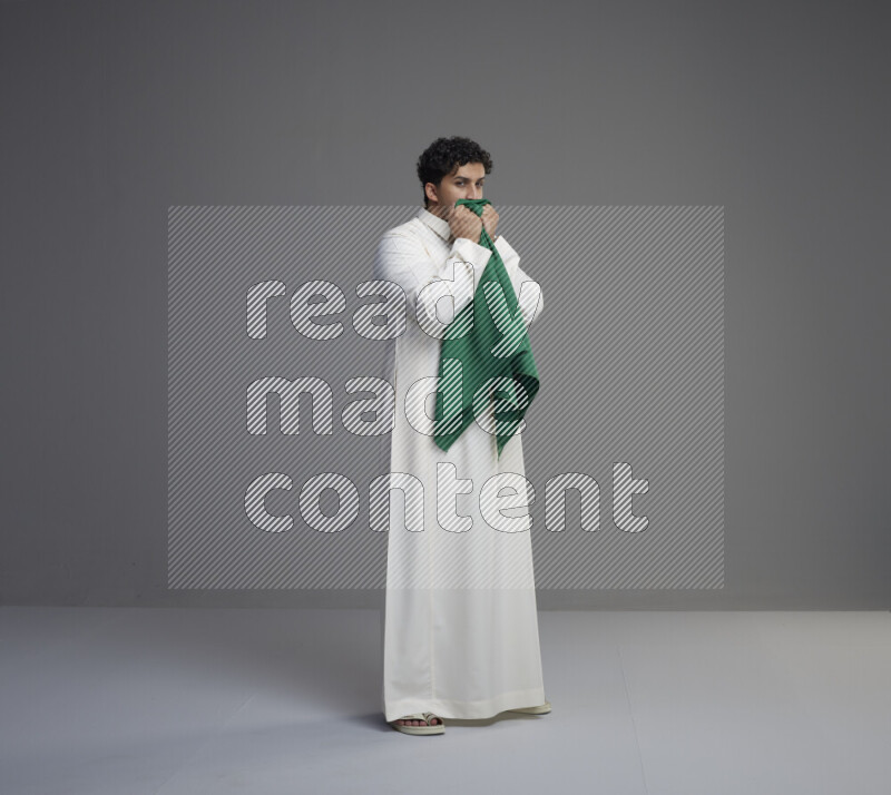 A Saudi man standing wearing thob kissing Saudi flag on gray background