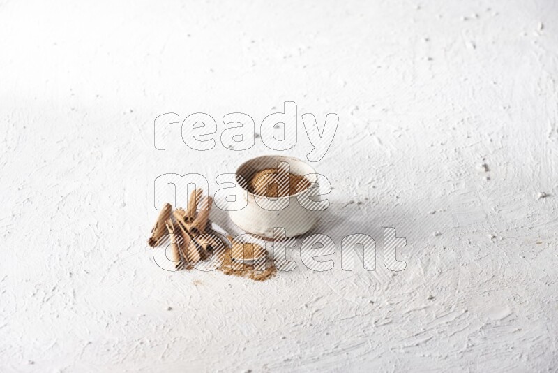 Ceramic beige bowl full of cinnamon powder and a metal spoon with cinnamon sticks next of it on a textured white background
