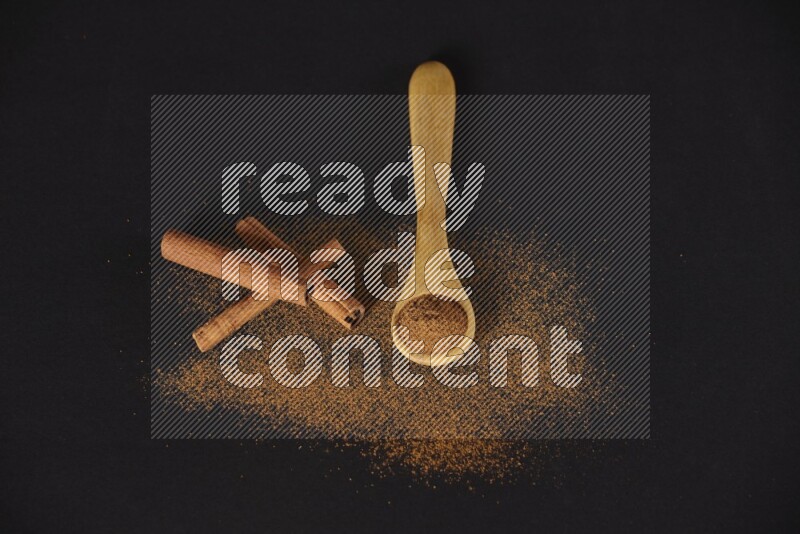Cinnamon powder in a wooden spoon and cinnamon sticks beside it on black background