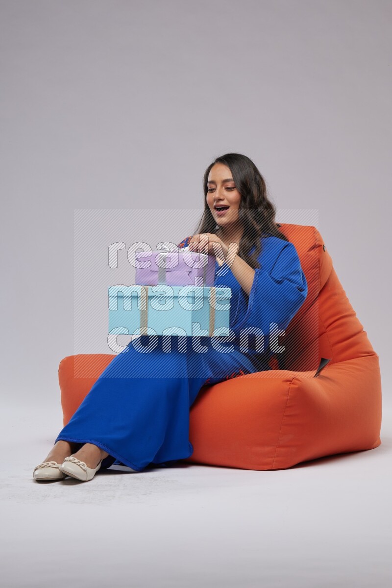 A woman sitting on an orange beanbag wearing Jalabeya holding a gift box