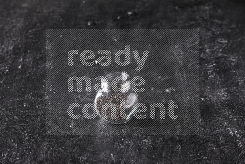 A glass spice jar full of black pepper beads on textured black flooring