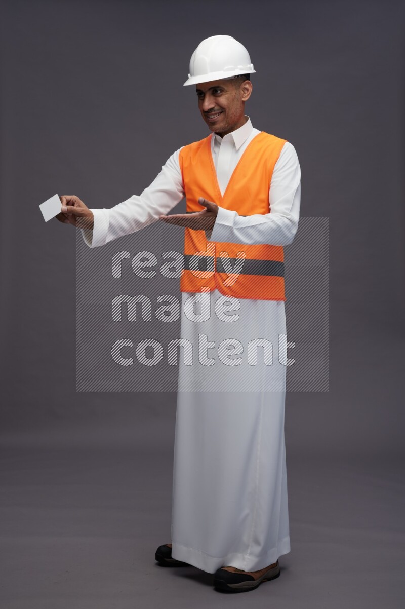 Saudi man wearing thob with engineer vest standing holding ATM card on gray background