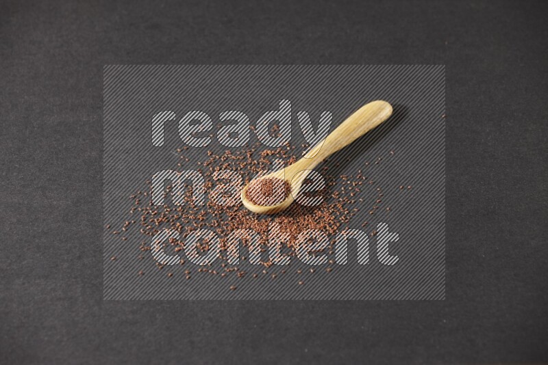 A wooden spoon full of garden cress seeds and seeds spread beside it on a black flooring