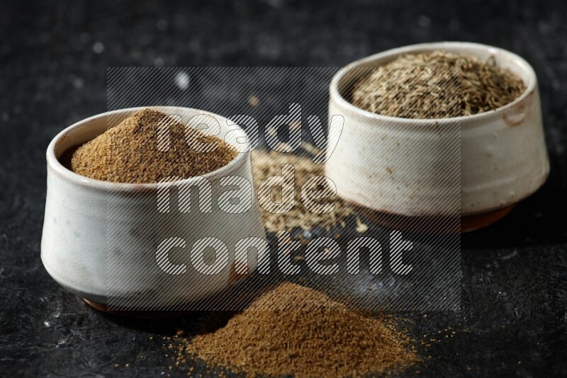2 beige bowls, one full of cumin powder and the other full of seeds on a textured black flooring