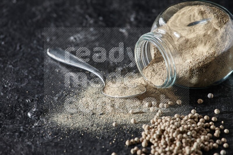 A flipped herbal glass jar and metal spoon full of white pepper powder with spilled powder and pepper beads on textured black flooring