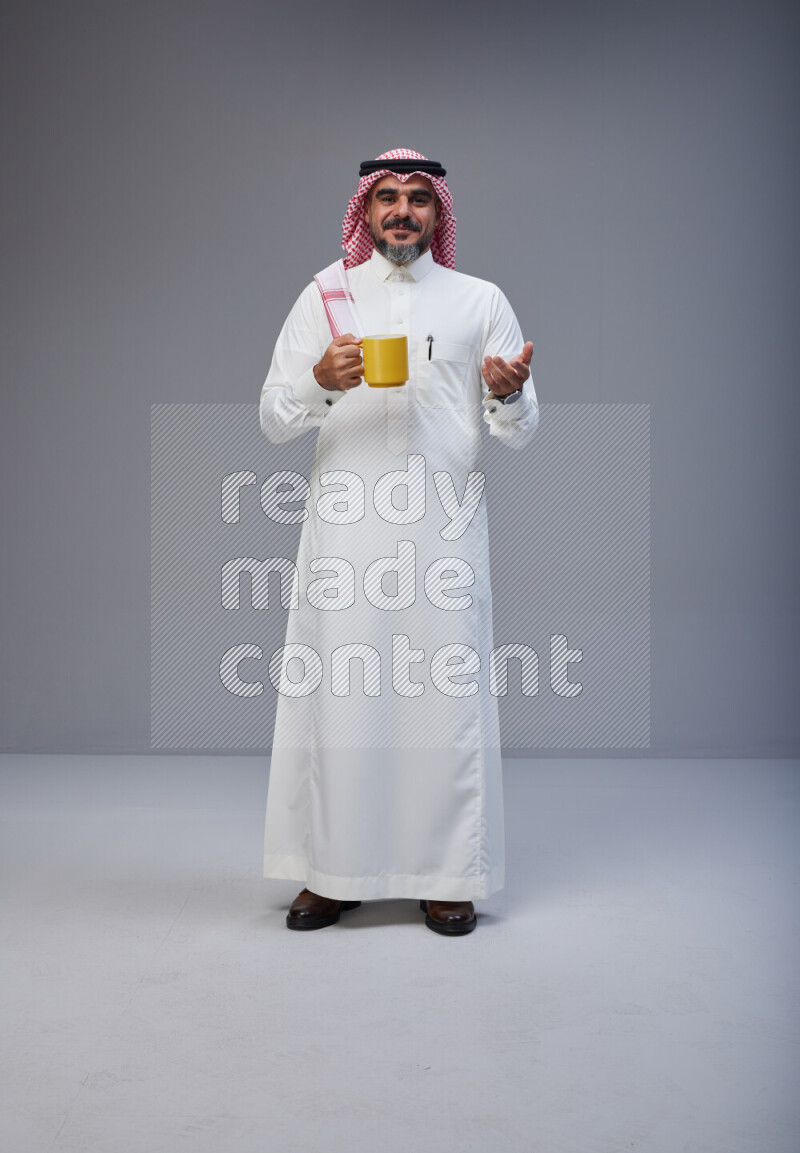 Saudi man Wearing Thob and red Shomag standing holding mug on Gray background