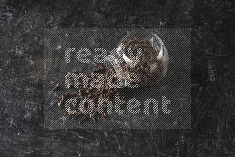 وعاء زجاجي للبهارات مقلوب ممتلئ بالقرنفل والقرنفل سقط منه على أرضية سوداء