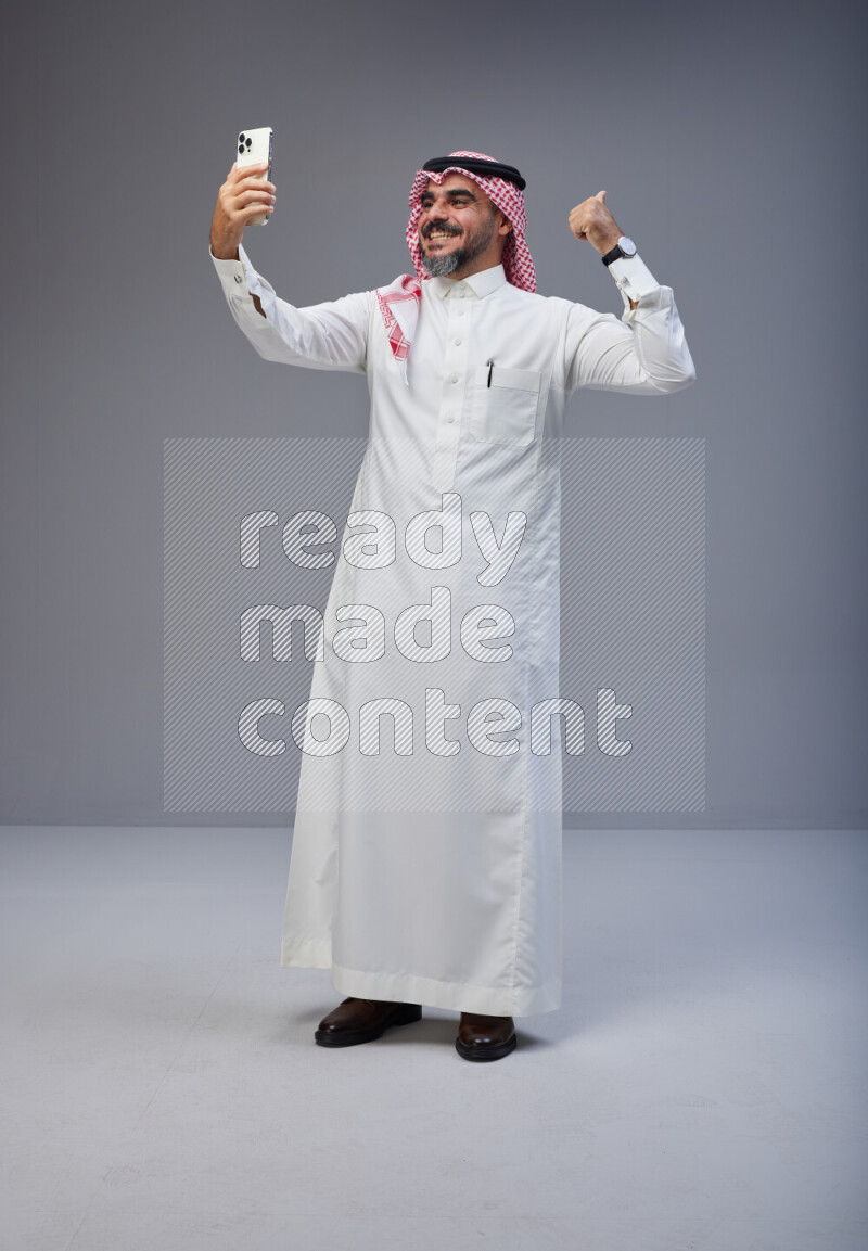 Saudi man Wearing Thob and red Shomag standing taking selfie on Gray background