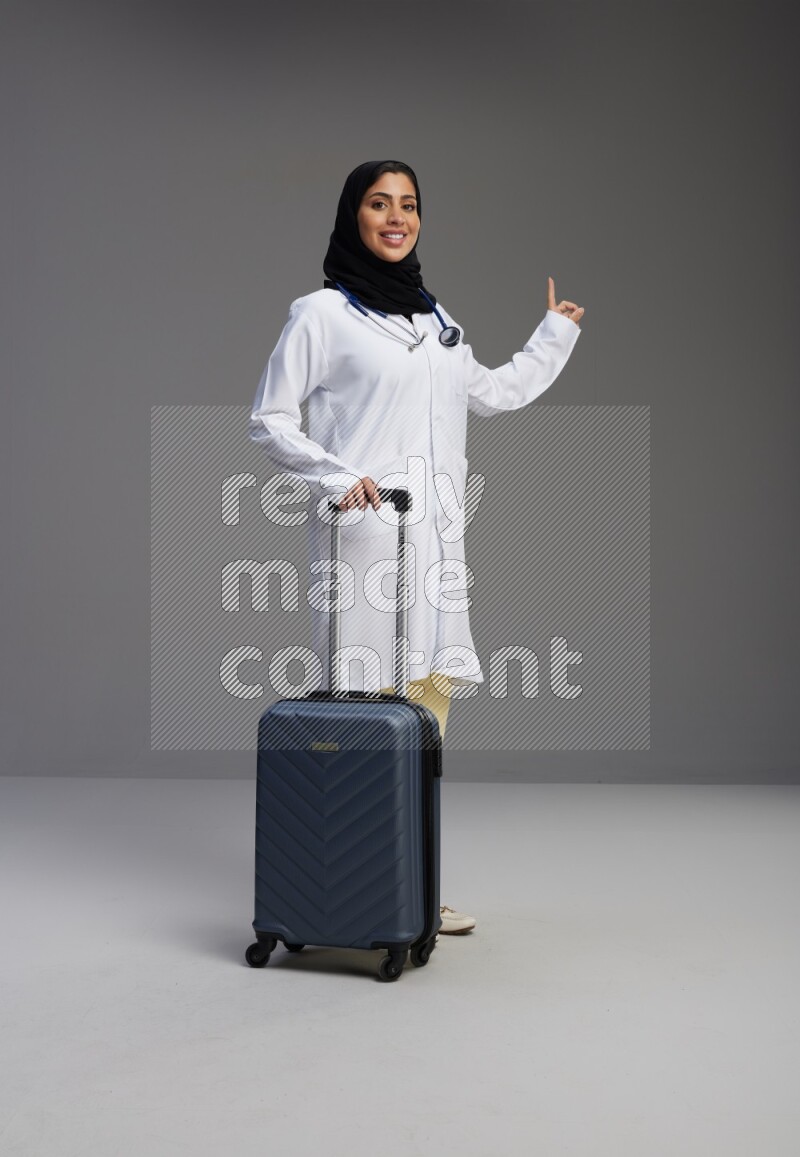 Saudi woman wearing lab coat with stethoscope standing holding Travel bag on Gray background