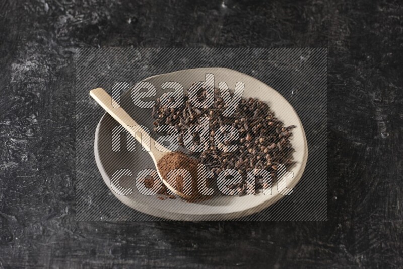 A Pottery plate full of cloves and a wooden spoon full of cloves powder on it on a textured black background