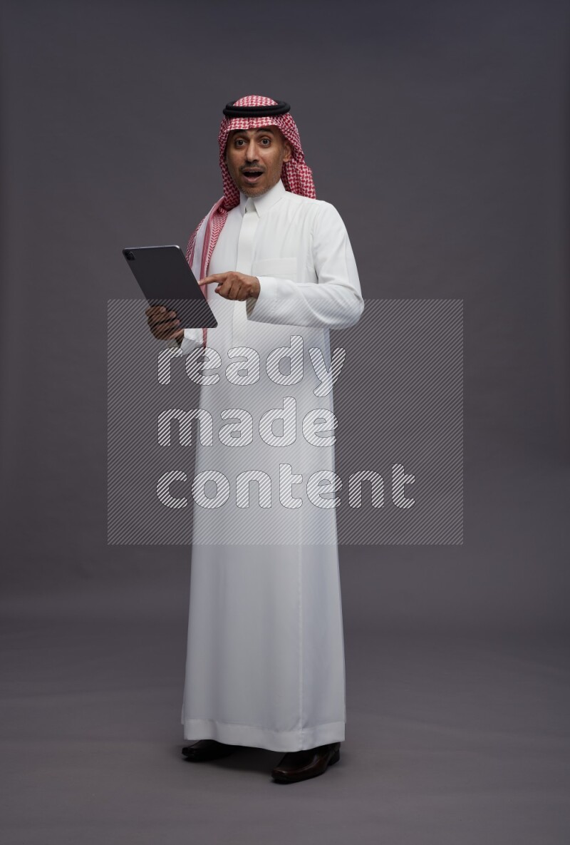 Saudi man wearing thob and shomag standing working on tablet on gray background