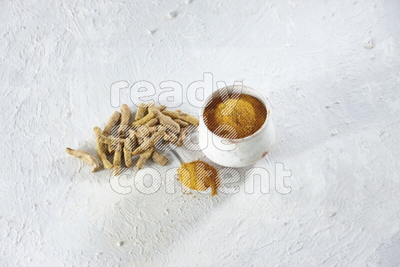 A beige pottery bowl and metal spoon full of turmeric powder and dried turmeric fingers next of them on textured white flooring