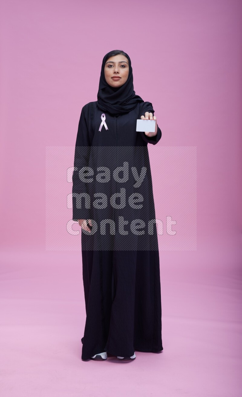Saudi woman wearing pink ribbon on Abaya standing holding ATM card on pink background