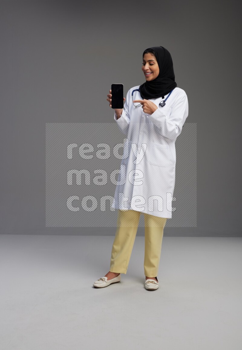 Saudi woman wearing lab coat with stethoscope standing showing phone to camera with sign in the back on Gray background