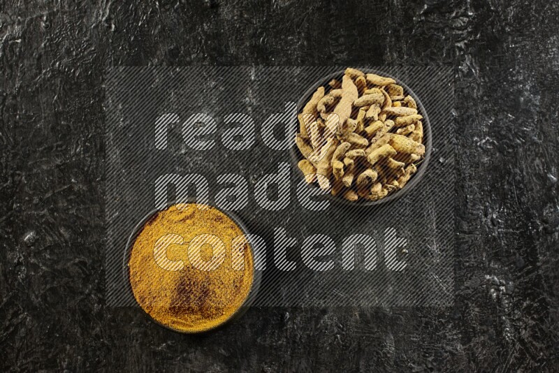 2 pottery black bowls, one full of turmeric powder and the other full of dried turmeric whole fingers on a textured black flooring