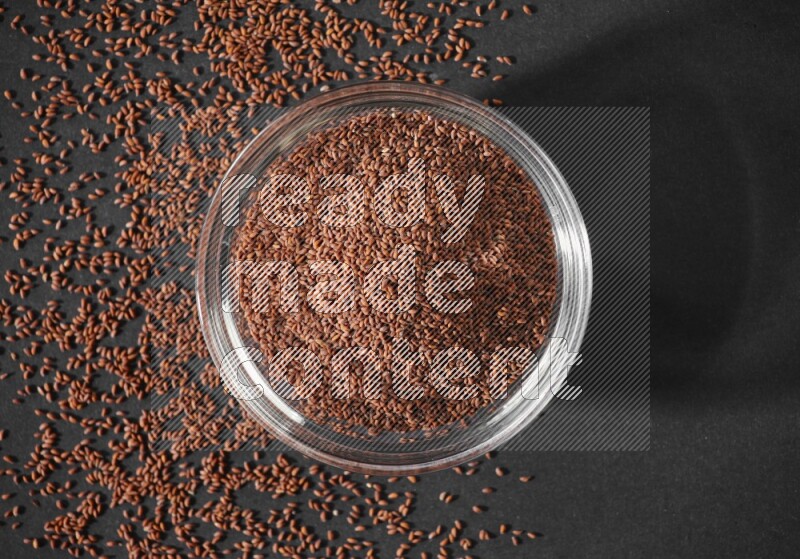A glass bowl full of garden cress seeds surrounded by seeds on a black flooring
