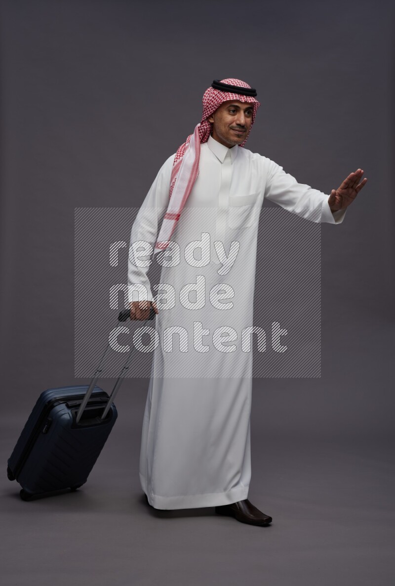 Saudi man wearing thob and shomag standing holding bag on gray background