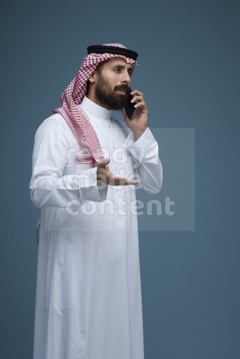 A man having a Call in a blue background wearing Saudi Thob with Shomag