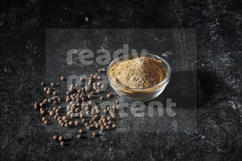 A glass bowl full of allspice powder and whole balls spreaded on a black flooring