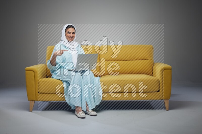 A Saudi woman sitting on a yellow Sofa With her Laptop Pointing wearing Blue Abaya