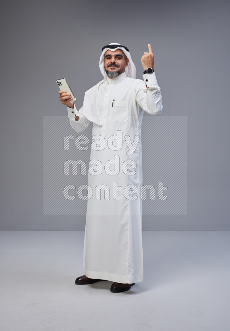Saudi man Wearing Thob and white Shomag standing texting on phone on Gray background
