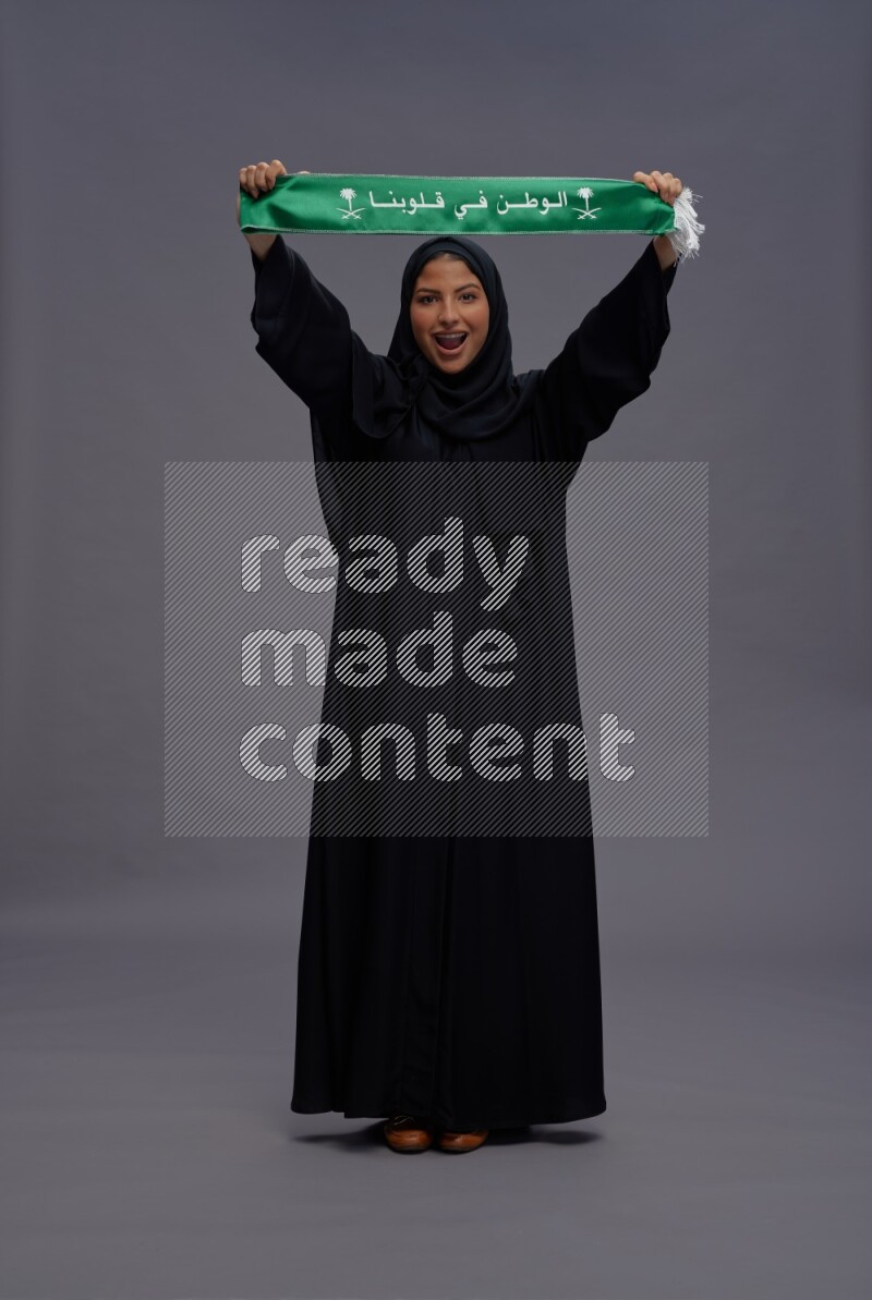 Saudi woman wearing Abaya standing holding Saudi flag on gray background