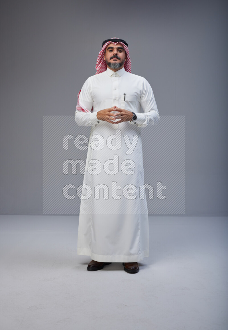 Saudi man Wearing Thob and red white Shomag standing interacting with the camera on Gray background