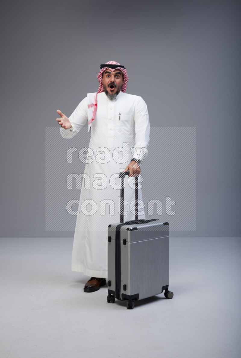 Saudi man wearing Thob and red Shomag standing holding Travel bag on Gray background