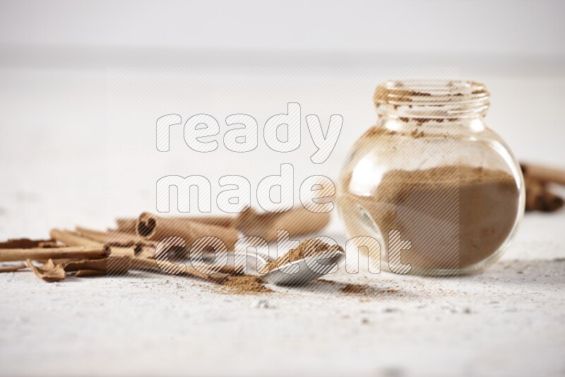 Herbal glass jar full cinnamon powder and a metal spoon surrounded by cinnamon sticks on a white background