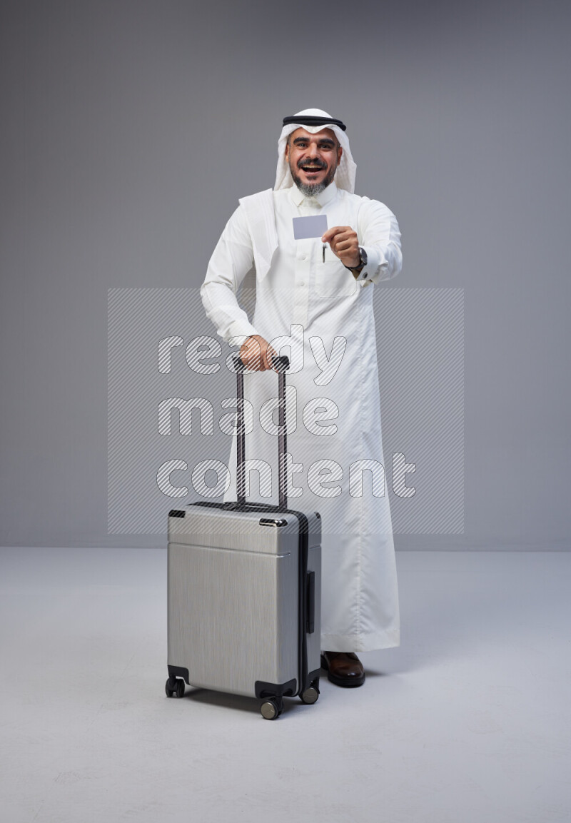Saudi man wearing Thob and white Shomag standing holding Travel bag and ATM card on Gray background