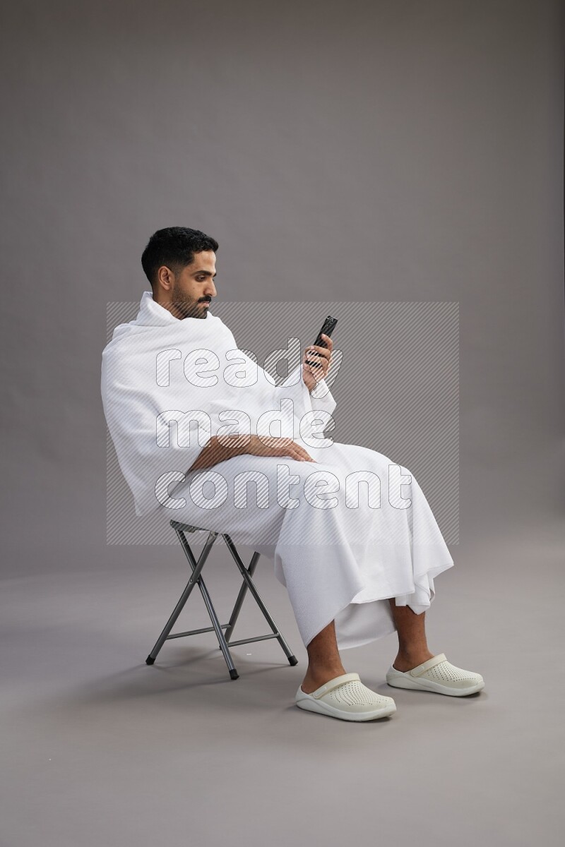A man wearing Ehram sitting on chair texting on phone on gray background