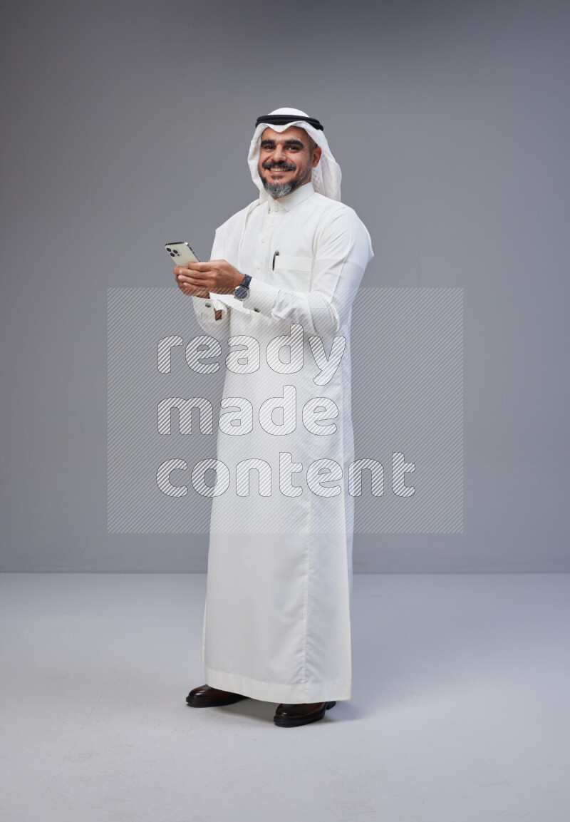 Saudi man Wearing Thob and white Shomag standing texting on phone on Gray background