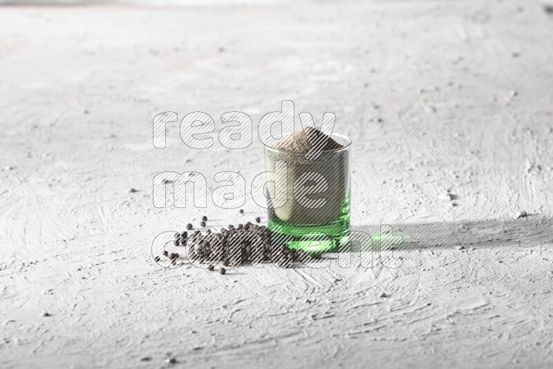 A glass cup full of black pepper powder and black pepper spread on a textured white flooring