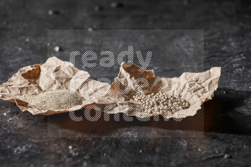 White pepper beads and powder in 2 crumpled paper on textured black flooring