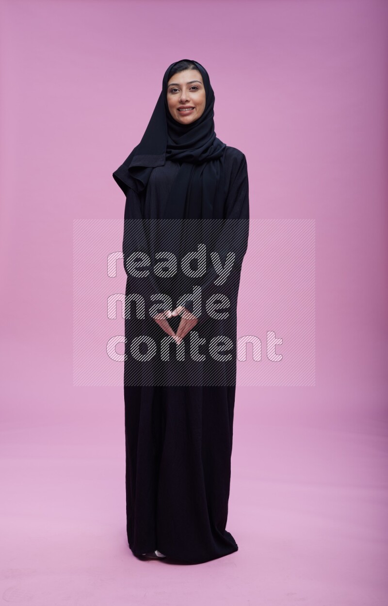 Saudi woman wearing Abaya standing interacting with the camera on pink background
