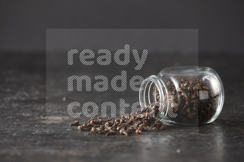 A flipped glass spice jar full of cloves and cloves came out of it on textured black flooring