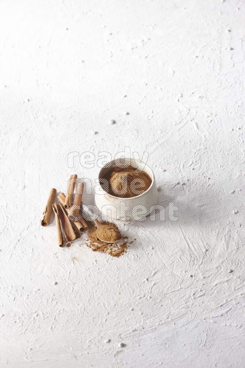 Ceramic beige bowl full of cinnamon powder and a metal spoon with cinnamon sticks next of it on a textured white background