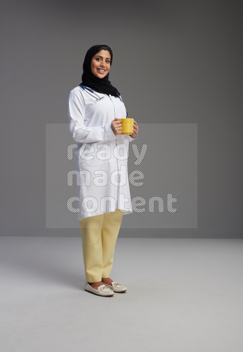 Saudi woman wearing lab coat with stethoscope standing holding mug on Gray background
